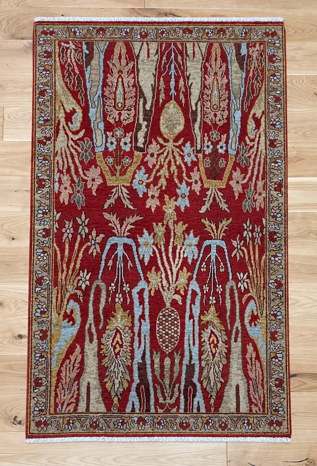 A rectangular area rug with a traditional ornate pattern in deep red, beige, light blue, and gold, laid flat on a light wood floor.
