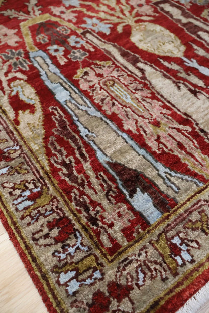 A close-up of a traditional patterned area rug with a deep red field, beige borders, and motifs in brown, cream, and light blue, laid out on a light wood floor.