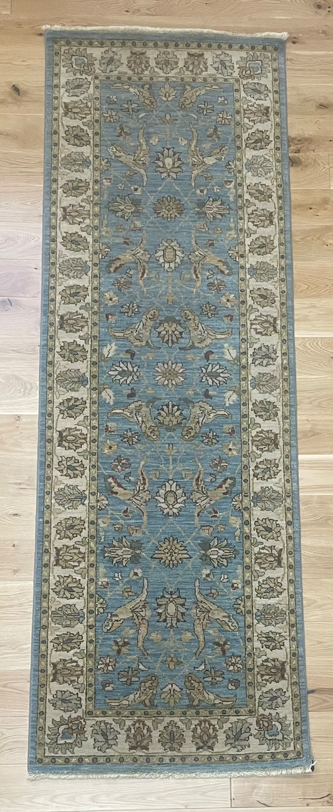 A rectangular area rug with a traditional floral pattern, featuring a dusty blue center field and beige borders, laid flat on a light wood floor.