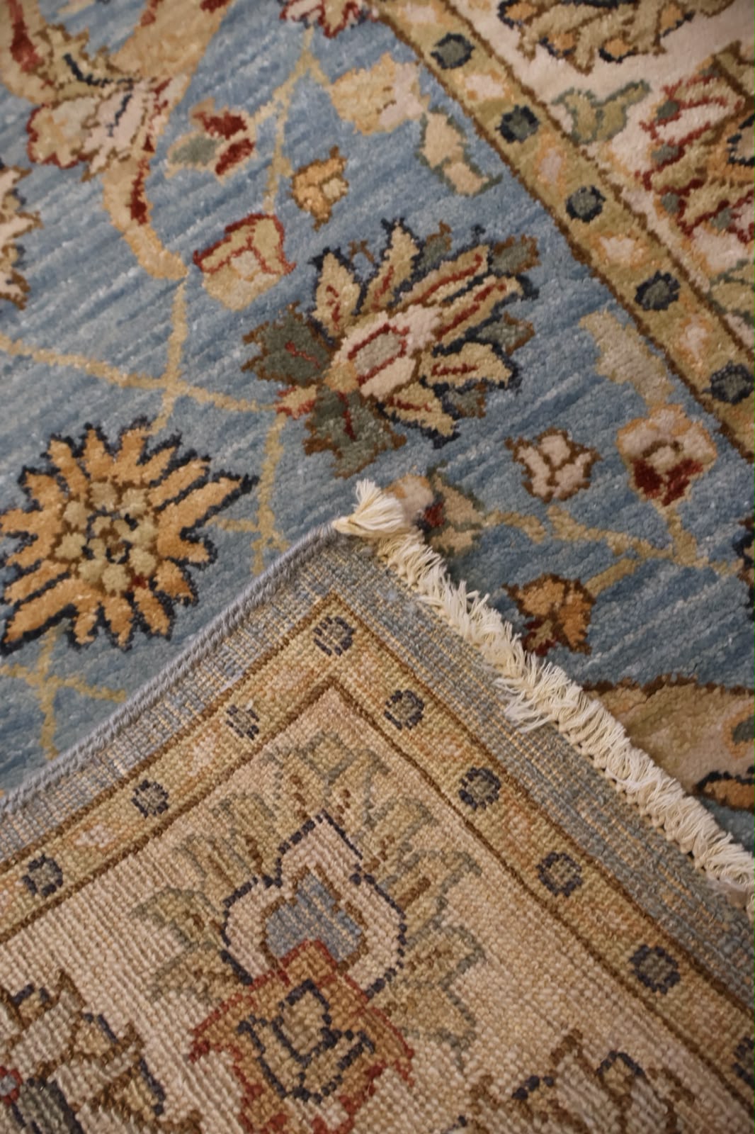 A close-up of a traditional patterned rug showing a light blue field with beige, brown, and rust floral motifs, meeting a beige border section with cream fringe.