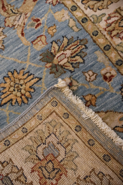 A close-up of a traditional patterned rug showing a light blue field with beige, brown, and rust floral motifs, meeting a beige border section with cream fringe.