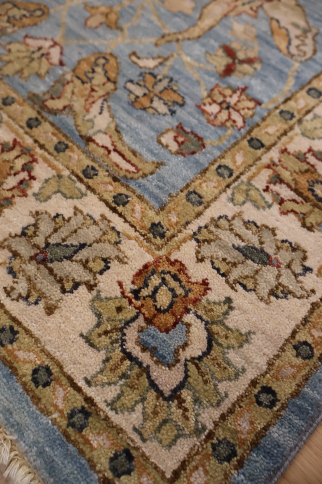 Close-up of a traditional patterned area rug corner showing a light blue field, beige border, and floral motifs in beige, rust, and muted green.