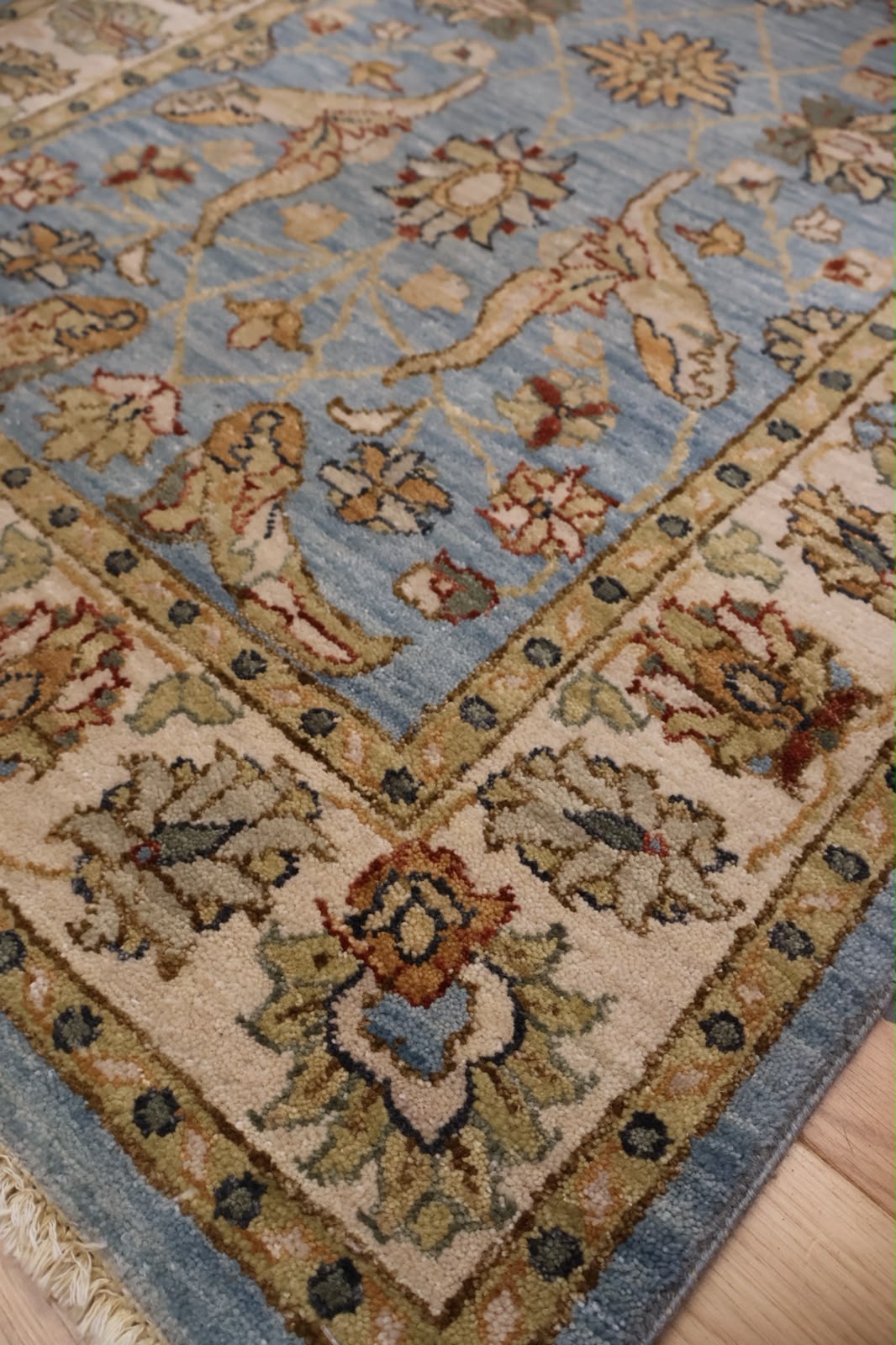 A close-up of a traditional patterned area rug with a dusty blue field, beige borders, and floral motifs in muted green, rust, and cream, laid over a light wood floor.