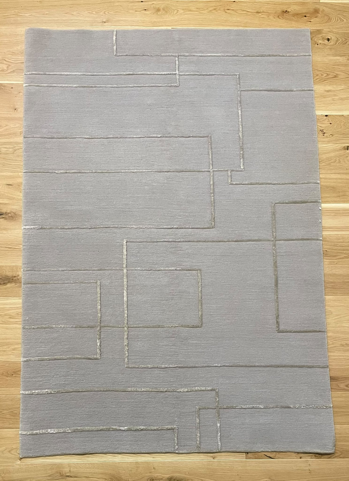 A rectangular light gray area rug with a geometric pattern of raised, darker gray lines, laid flat on a light wood floor.