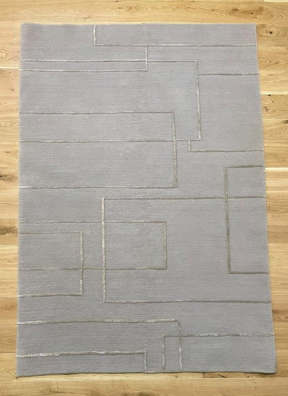 A rectangular light gray area rug with a geometric pattern of raised, darker gray lines, laid flat on a light wood floor.