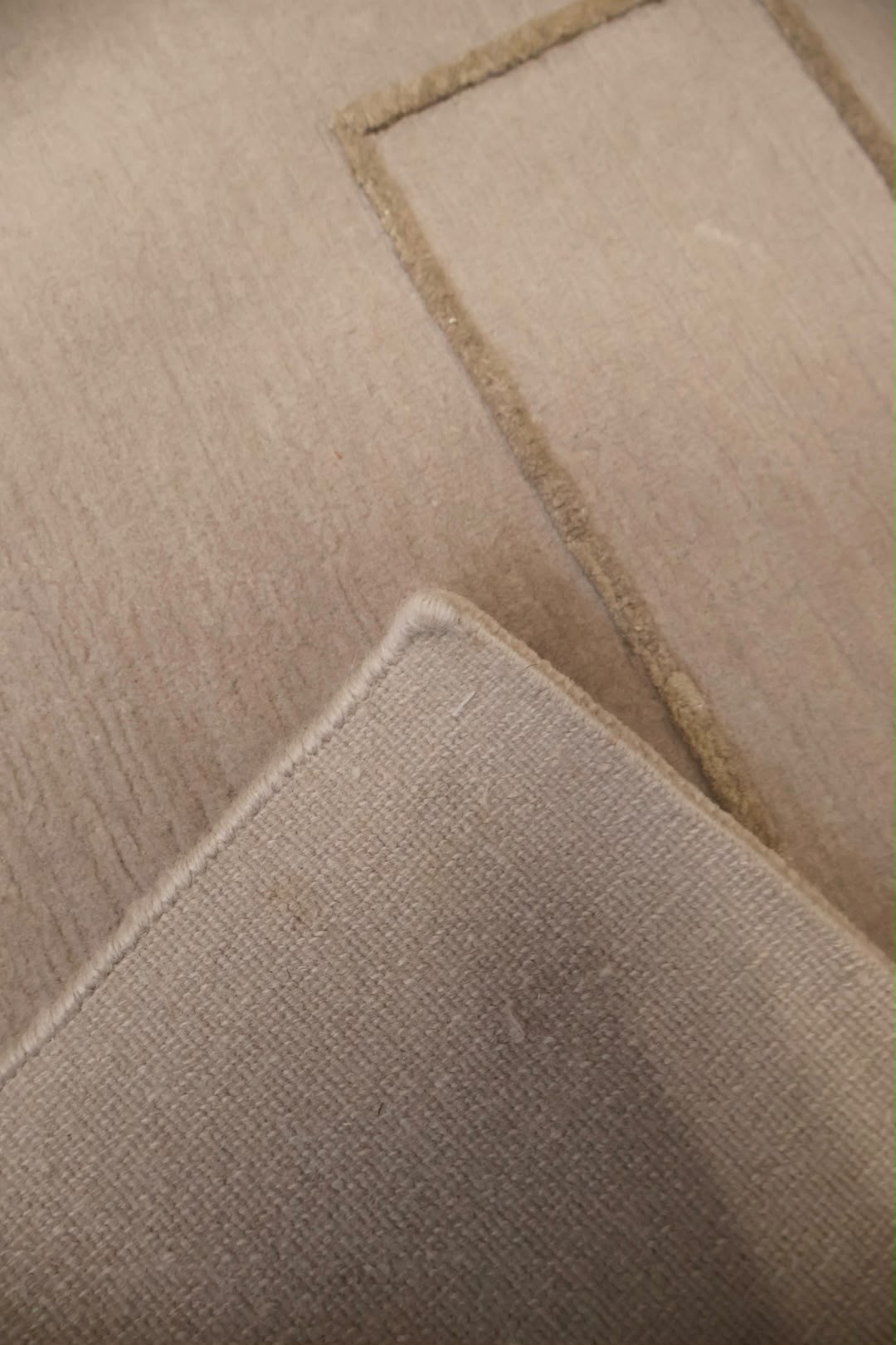 A close-up of a beige woven fabric with a stitched rectangular border and a folded corner showing the texture.