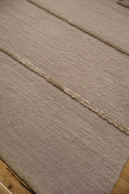 A close-up of a textured area rug in a warm taupe-beige color, featuring a central raised stripe with a braided or tufted edge detail, laid over a light wood floor.