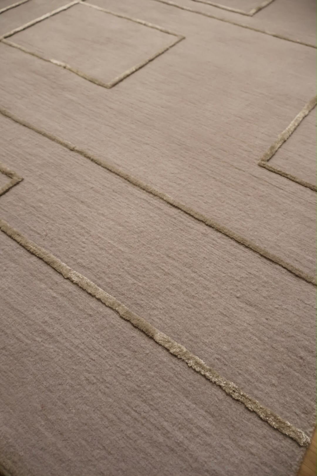 A close-up view of a textured area rug in a warm taupe-beige color, featuring a raised geometric pattern of concentric squares and rectangles outlined in a slightly lighter beige tone.