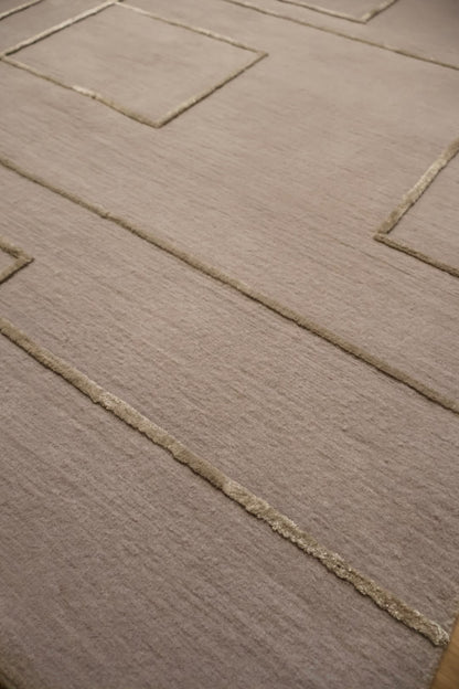 A close-up view of a textured area rug in a warm taupe-beige color, featuring a raised geometric pattern of concentric squares and rectangles outlined in a slightly lighter beige tone.
