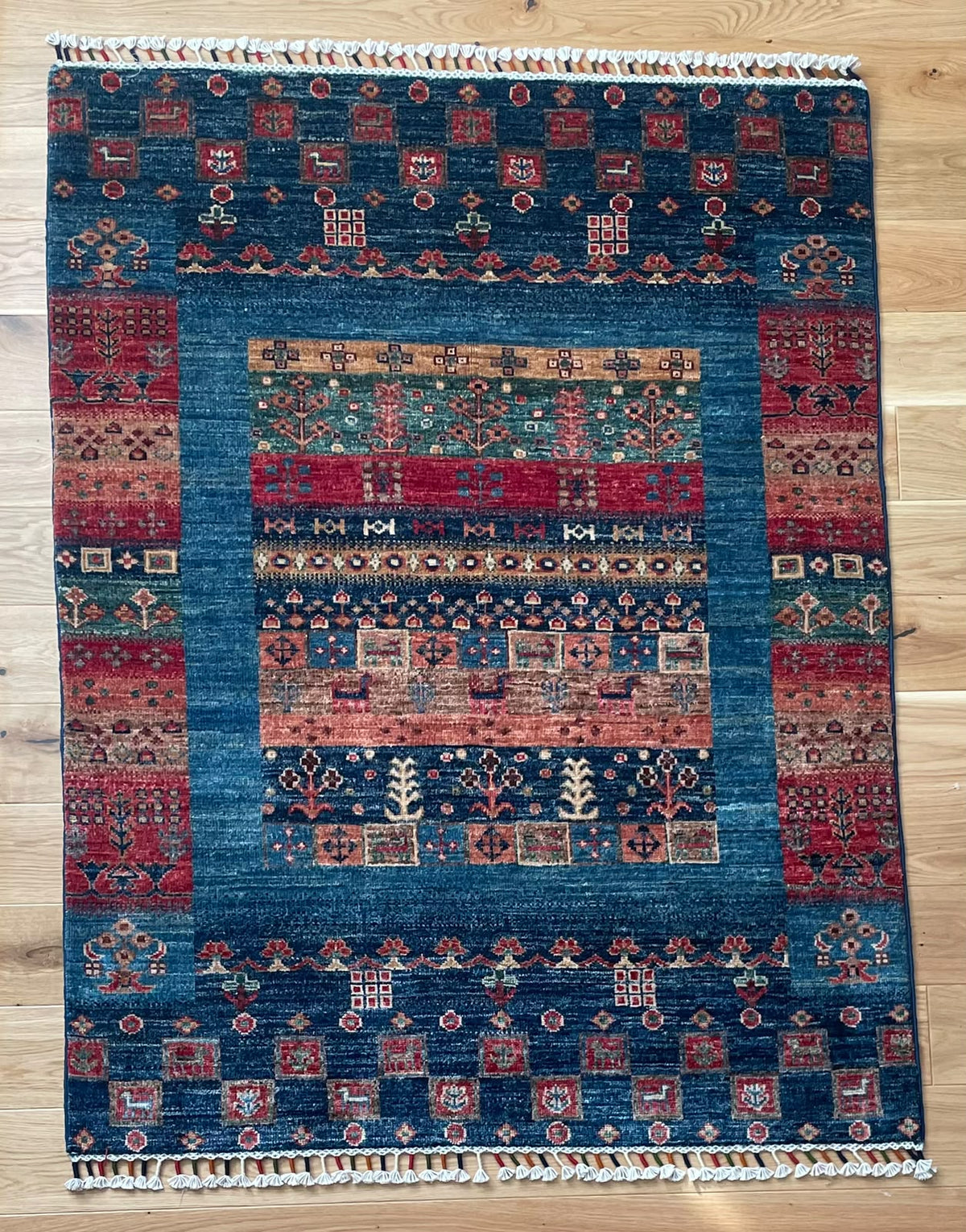 A rectangular hand-woven area rug with a traditional geometric pattern, featuring a dominant blue field with red, beige, and green accents, laid flat on a light wood floor.