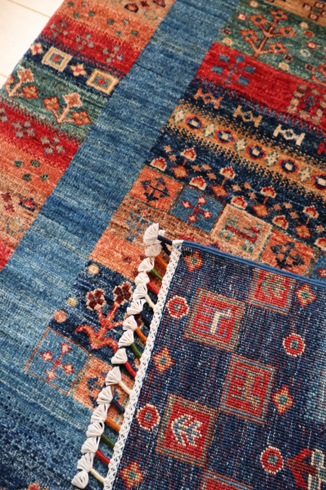 A close-up of a patterned area rug with a blue border and a multicolor geometric design in red, orange, and navy, featuring white tassels along one edge.