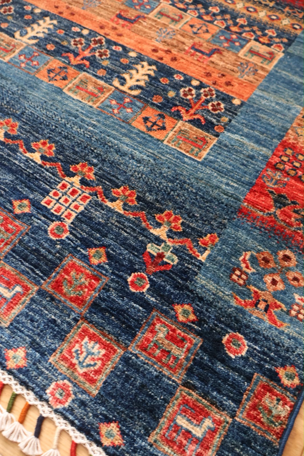 A close-up of a patterned area rug with a deep blue field, red and orange geometric motifs, and white fringe along the edge.