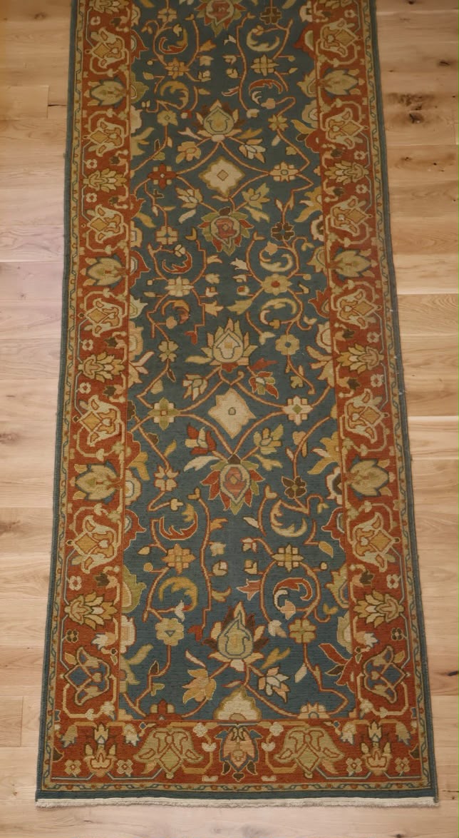 A traditional long runner rug with an intricate floral pattern, featuring a dark teal-blue center field, rust-red borders, and beige accents, laid out on a light wood floor.