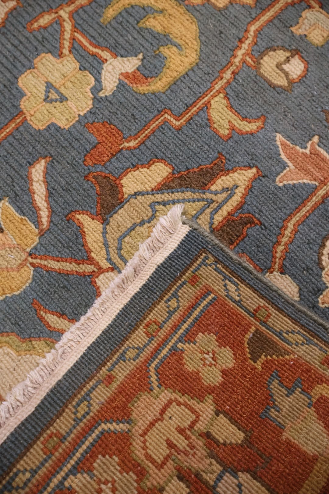 Close-up of a patterned area rug corner showing a dusty blue field with cream, rust, and brown floral motifs, plus a fringed edge and a rust-toned inner border.