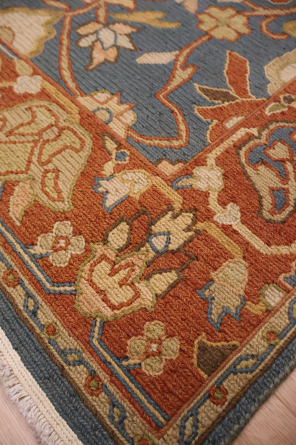 Close-up of a traditional patterned area rug with a rust orange field, blue border, and cream floral motifs, shown on a light wood floor.
