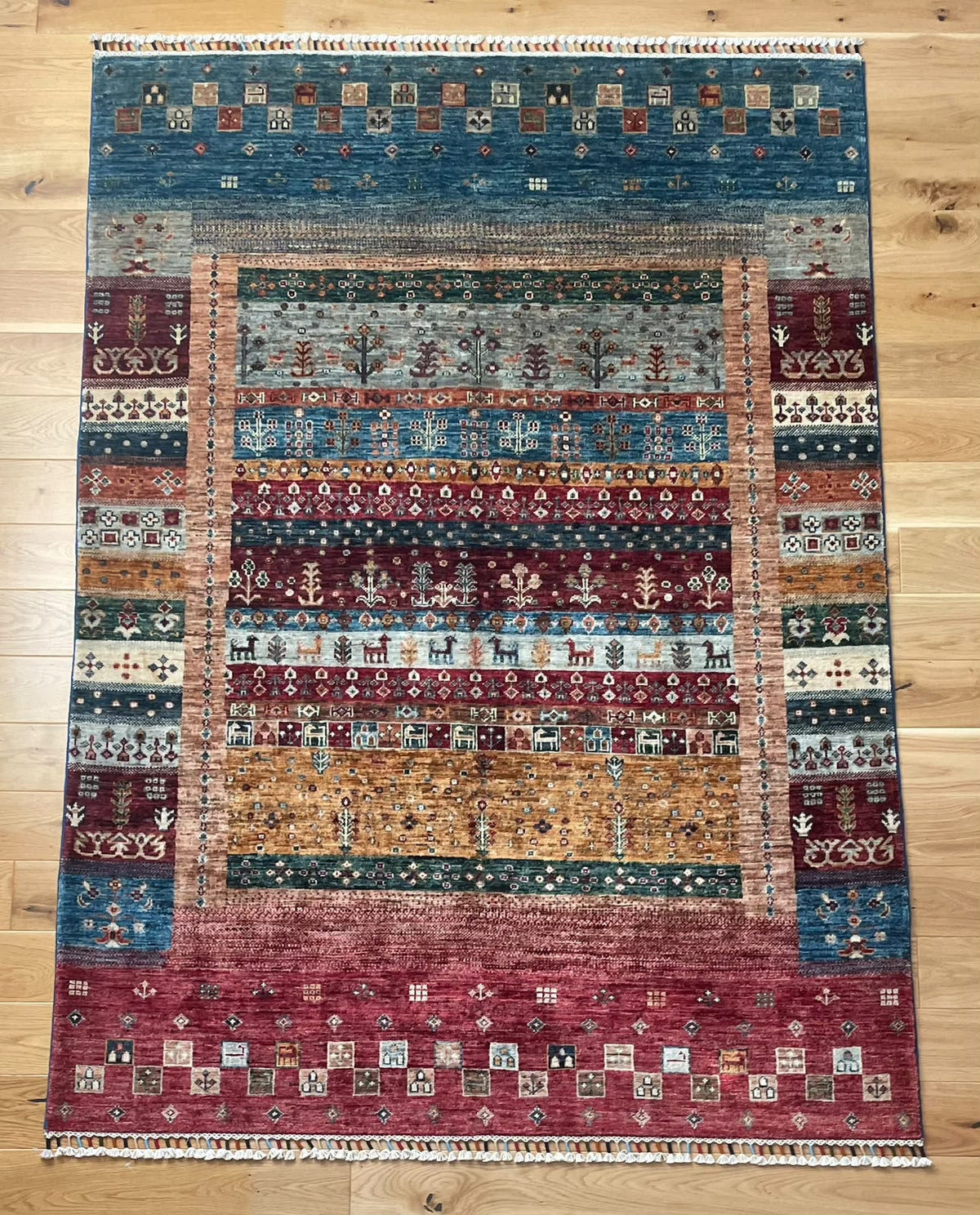 A rectangular hand-woven area rug with a complex geometric and tribal pattern, displayed flat on a light wood floor. The rug features a multi-colored design with prominent bands of blue, burgundy red, rust orange, beige, and green, finished with a fringed border.