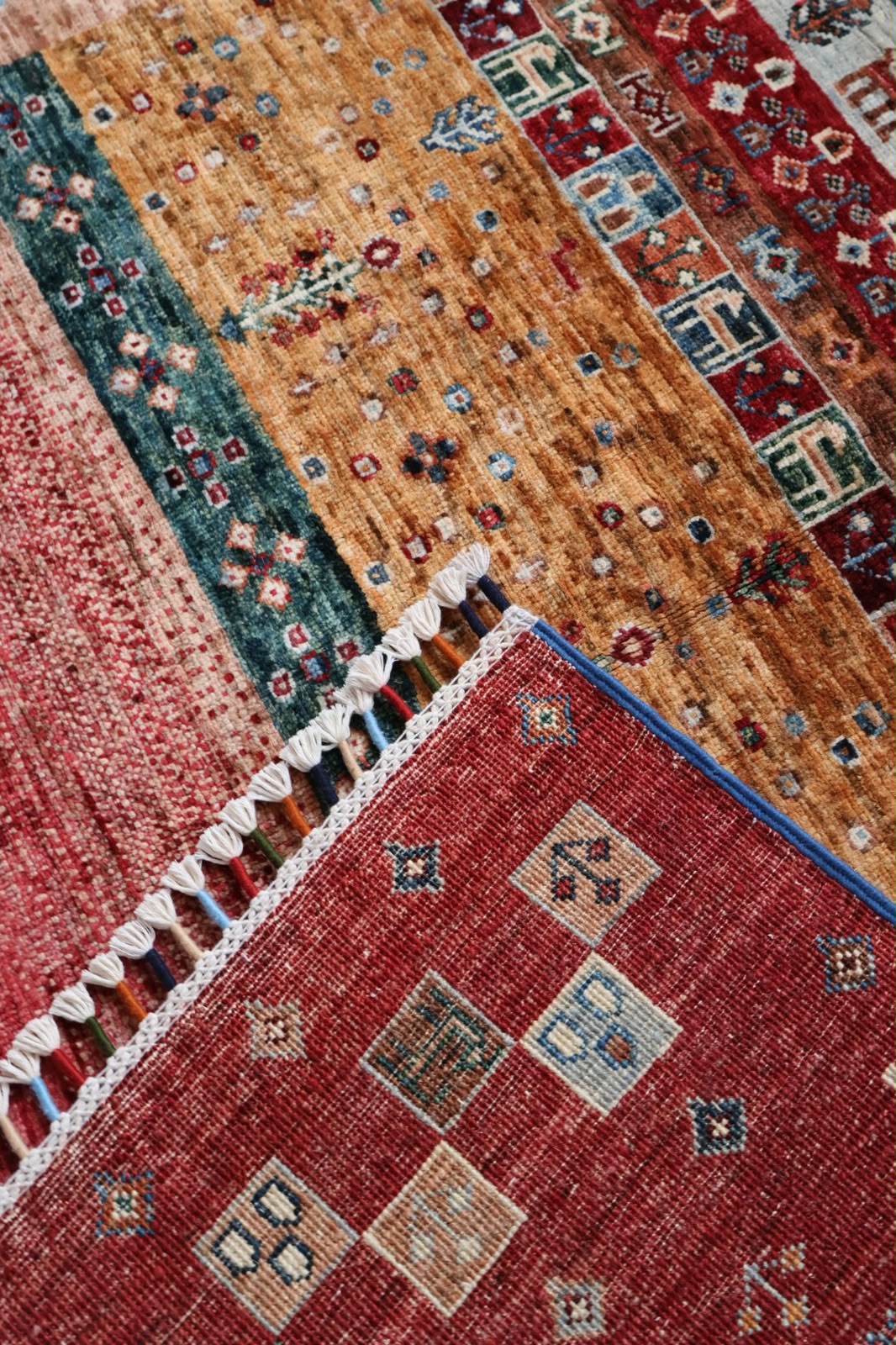 A close-up of a multi-patterned area rug showing different sections with distinct designs, including a prominent red section with beige geometric motifs, a teal patterned section, and a tan section with small floral details.