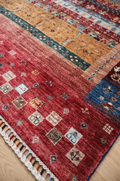 A close-up of a traditional patterned area rug with a red field, geometric motifs, and a white tassel fringe, laid on a light wood floor.