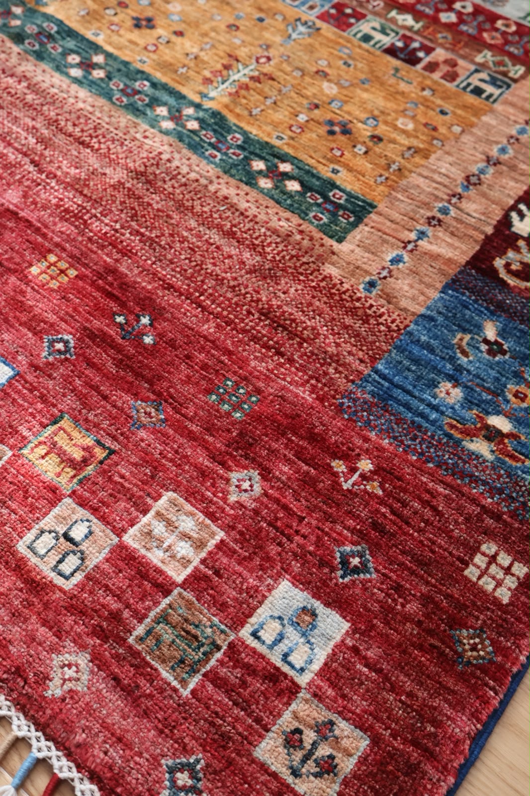 A close-up of a handwoven area rug with a rustic patchwork design, featuring a dominant red field with geometric motifs, a blue section, and a warm orange section with floral patterns.