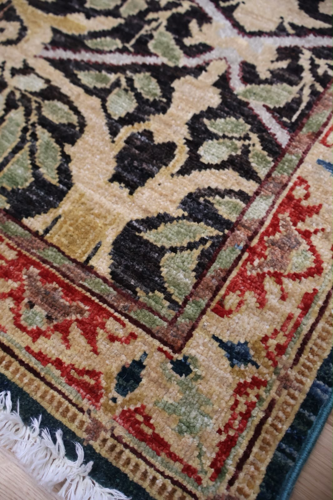 Close-up of a patterned area rug corner showing a cream base with dark brown and olive green floral motifs and a red border, with white fringe visible at the bottom.