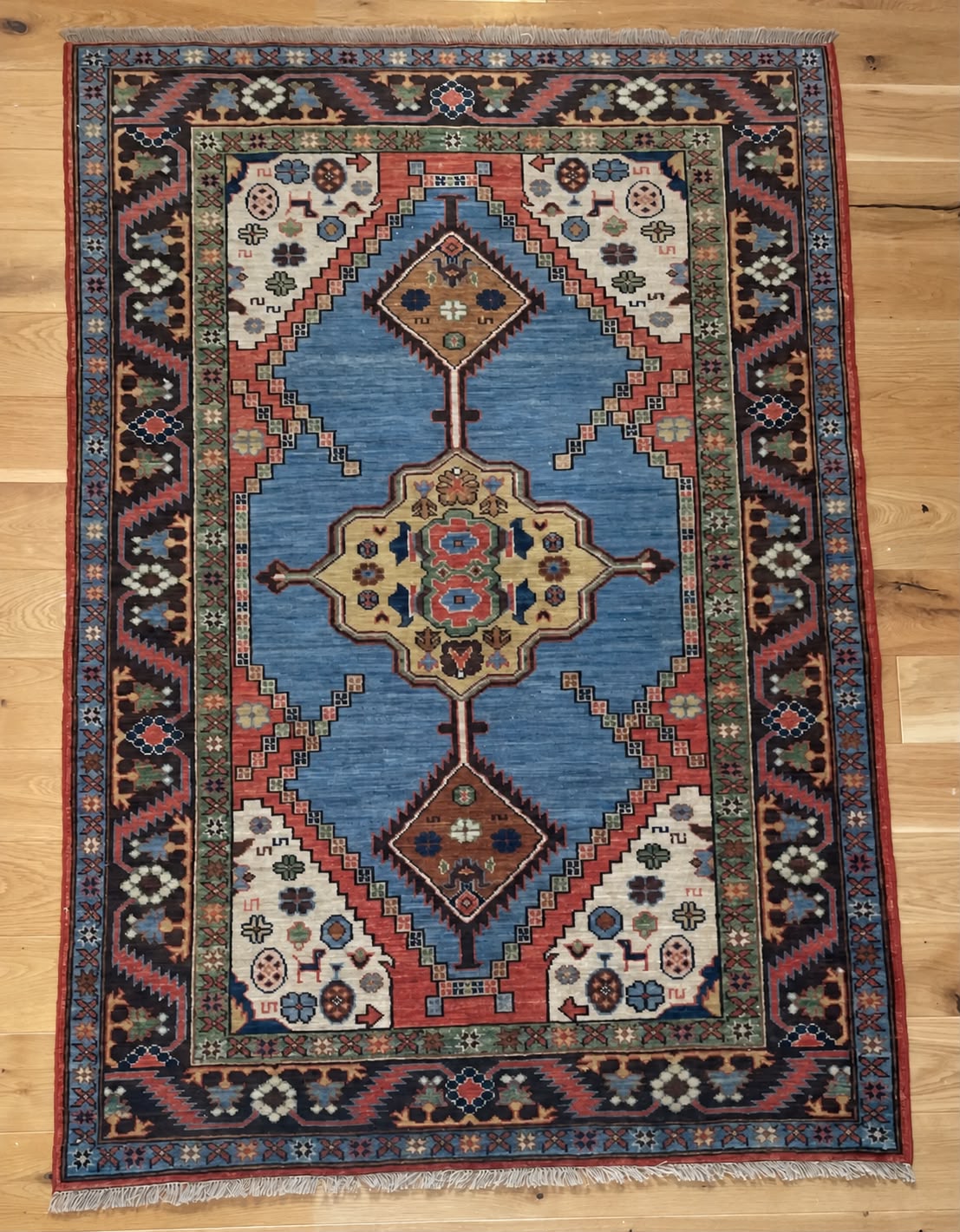 A traditional rectangular area rug with a complex geometric pattern, featuring a central blue field, beige and red medallions, and a dark border, laid flat on a light wood floor.