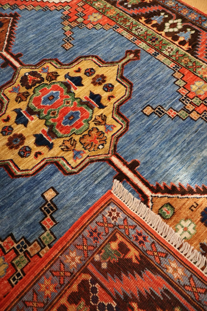 A close-up of a handwoven Persian-style rug featuring a large blue field with a central beige medallion decorated with red, green, and blue floral motifs, bordered by orange and brown geometric patterns with white fringe.