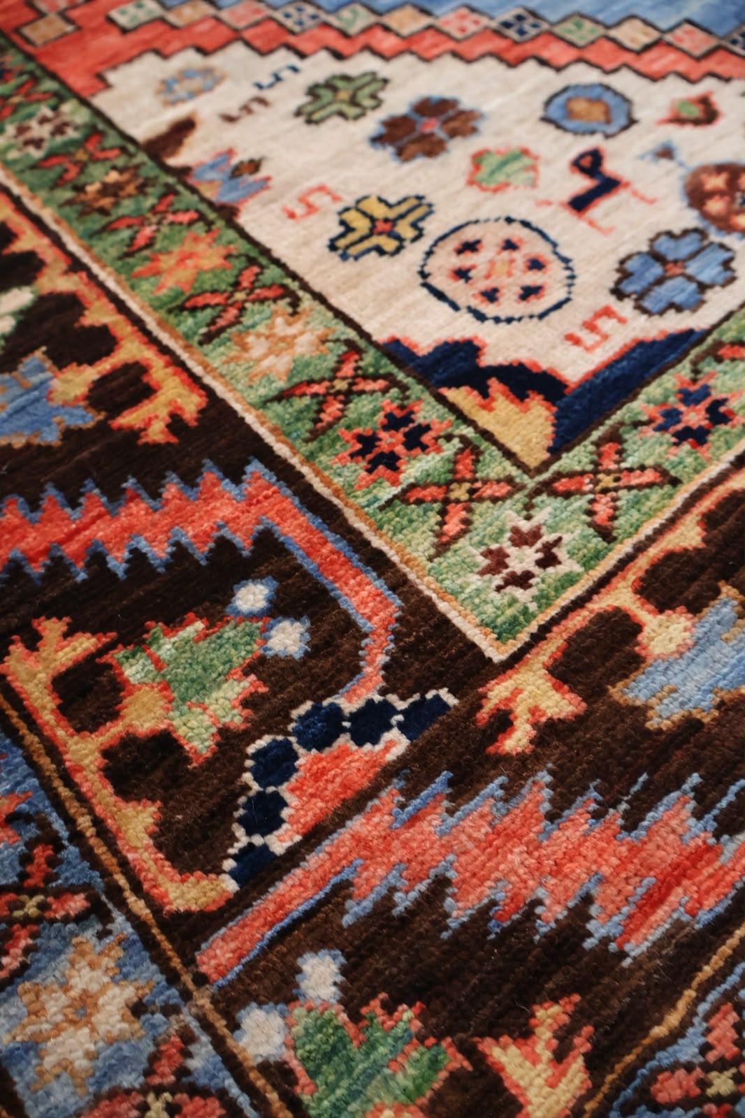 Close-up of a traditional patterned rug with geometric borders and floral motifs in dark brown, rust orange, cream, blue, and green.