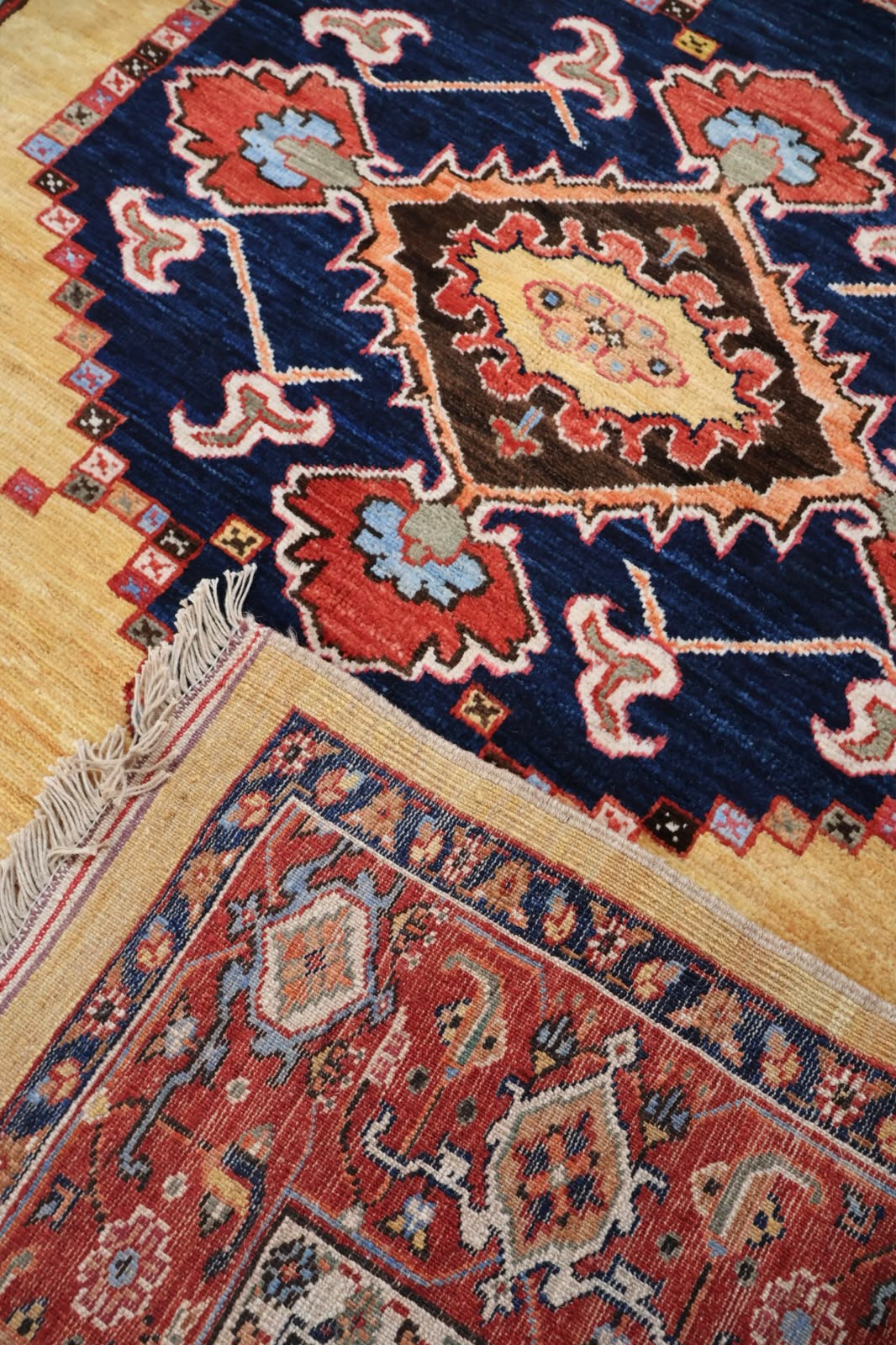 A close-up of a traditional patterned rug with a deep blue field, red and beige medallions, and cream fringe, shown alongside another red patterned rug on a light wood floor.