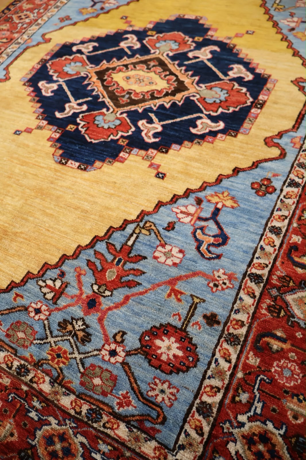 A close-up of a traditional patterned rug with a large mustard yellow field, a central navy blue medallion with red and beige details, and a light blue section with floral motifs and a red border.