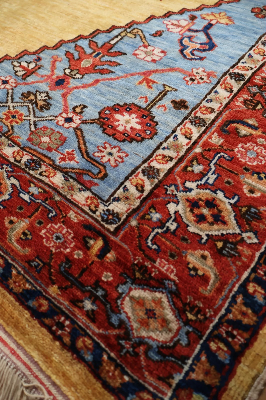 A close-up of a traditional patterned area rug showing its corner, featuring a light blue field, a red border, and cream floral motifs.