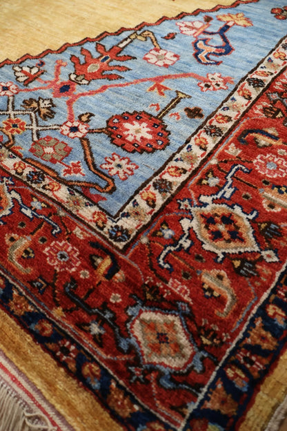 A close-up of a traditional patterned area rug showing its corner, featuring a light blue field, a red border, and cream floral motifs.