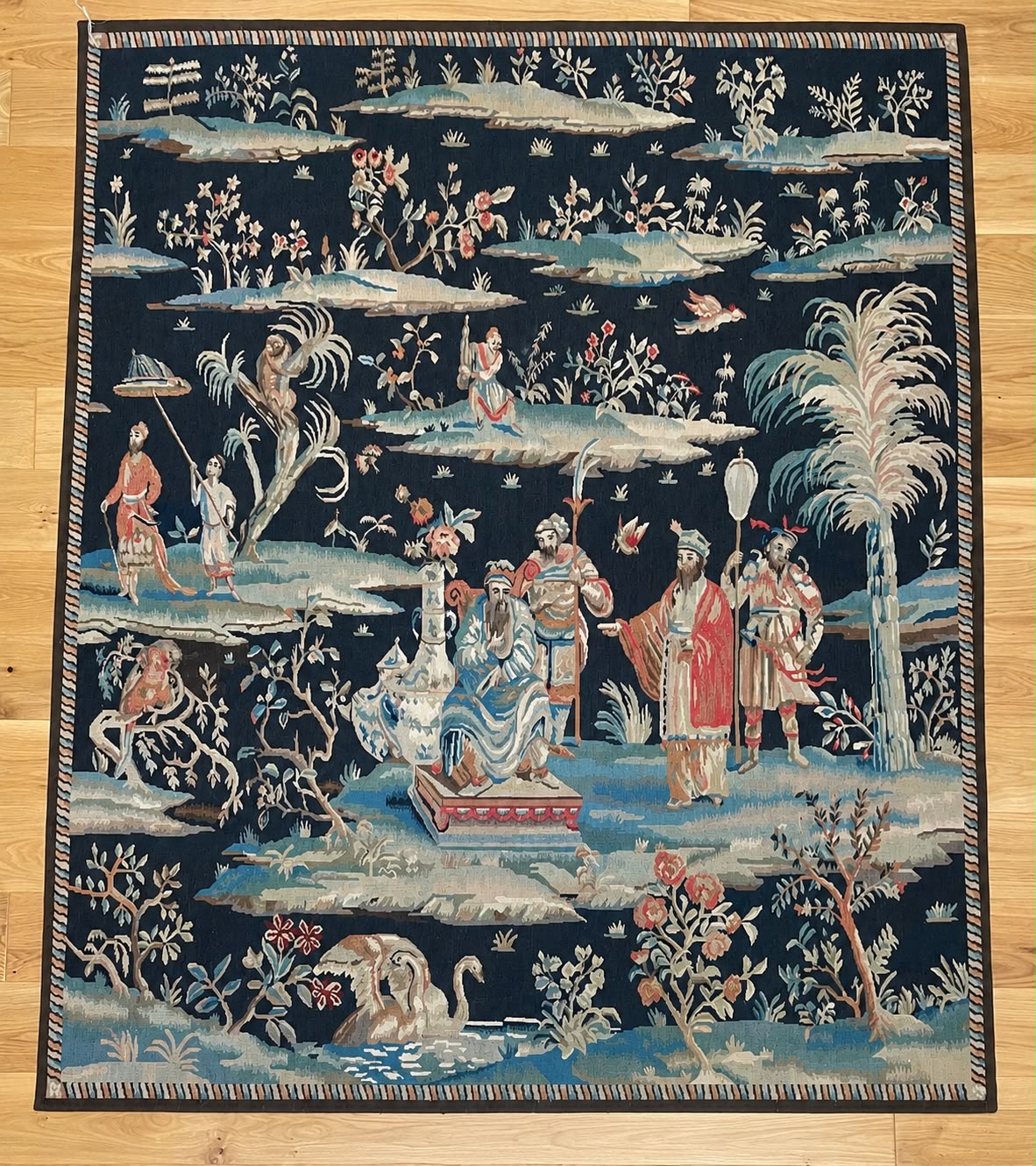 A square area rug with a dark navy blue background and an intricate, multi-colored chinoiserie scene featuring figures, palm trees, and floral motifs, laid flat on a light wood floor.