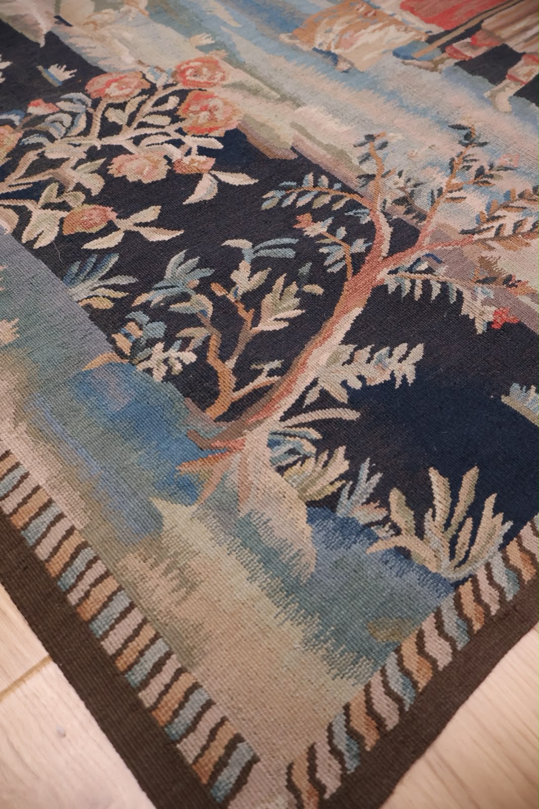 A close-up of a woven tapestry showing a detailed landscape scene with a tree, flowers, and a striped border, laid out on a light-colored wooden floor.