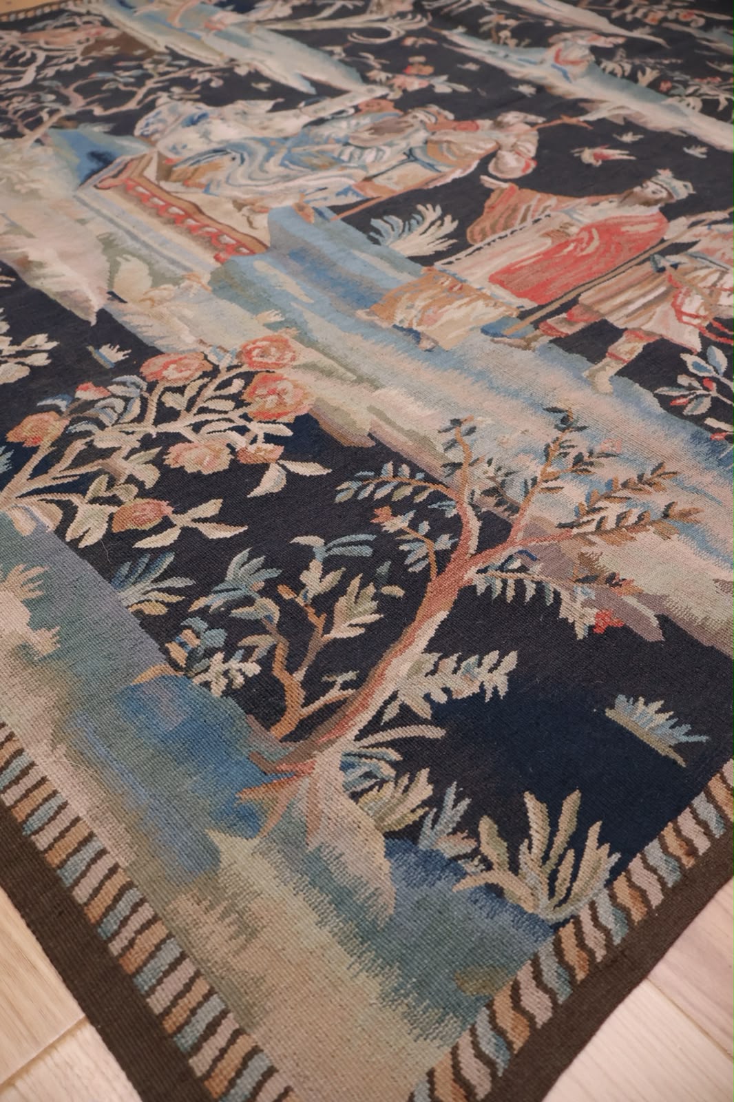 A close-up of an antique-style tapestry featuring a detailed landscape scene with figures, trees, and floral motifs in dark navy blue, beige, light blue, and rust red.