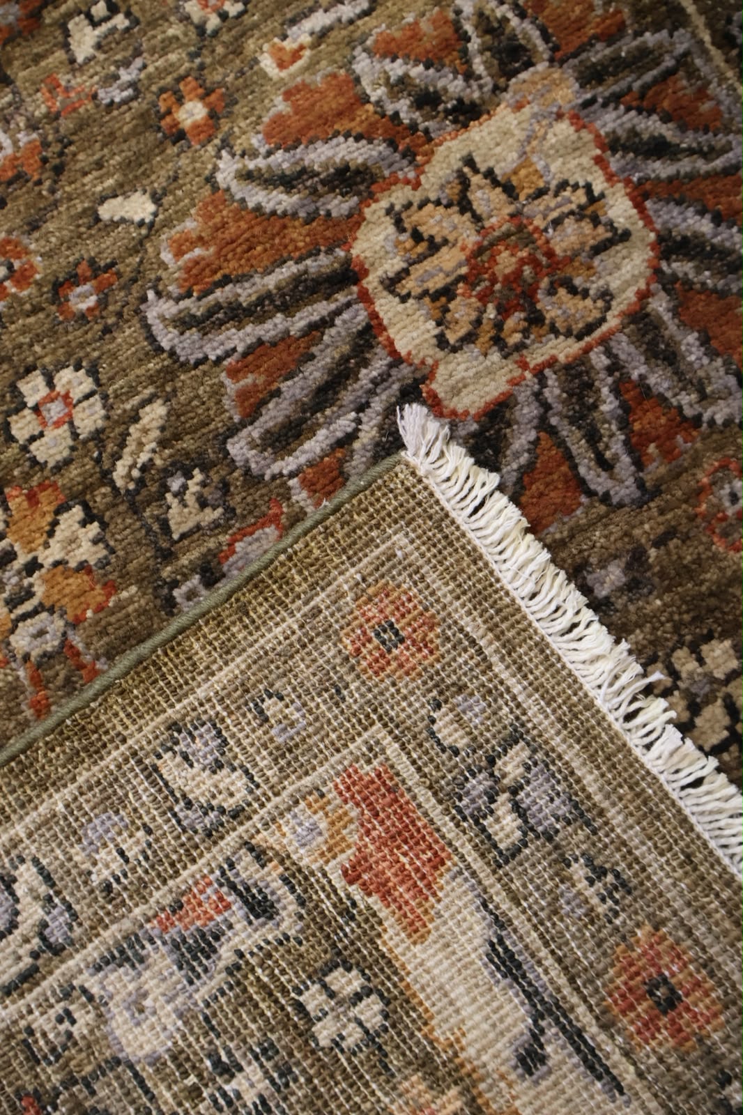 A close-up of a traditional patterned area rug showing its corner with white fringe, featuring a floral motif in earthy tones of brown, rust, beige, and gray.