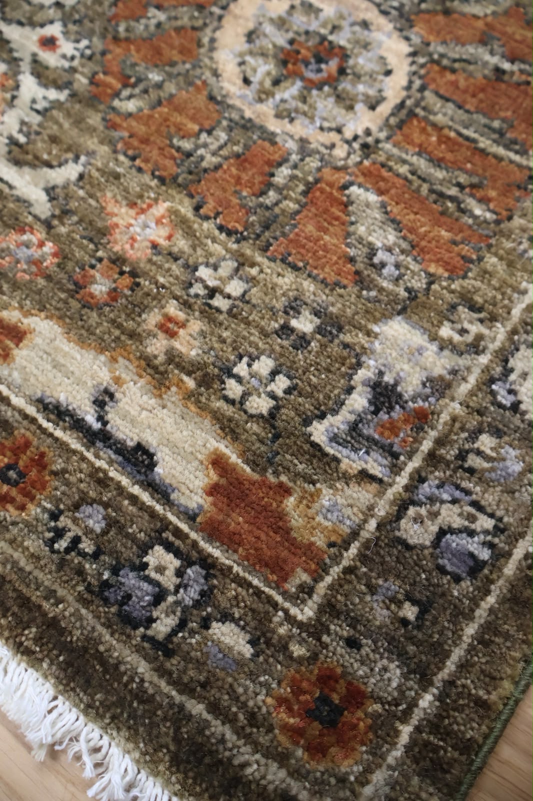 A close-up of a traditional patterned area rug with white fringe, featuring a complex design in brown, rust orange, beige, and muted blue-gray tones.