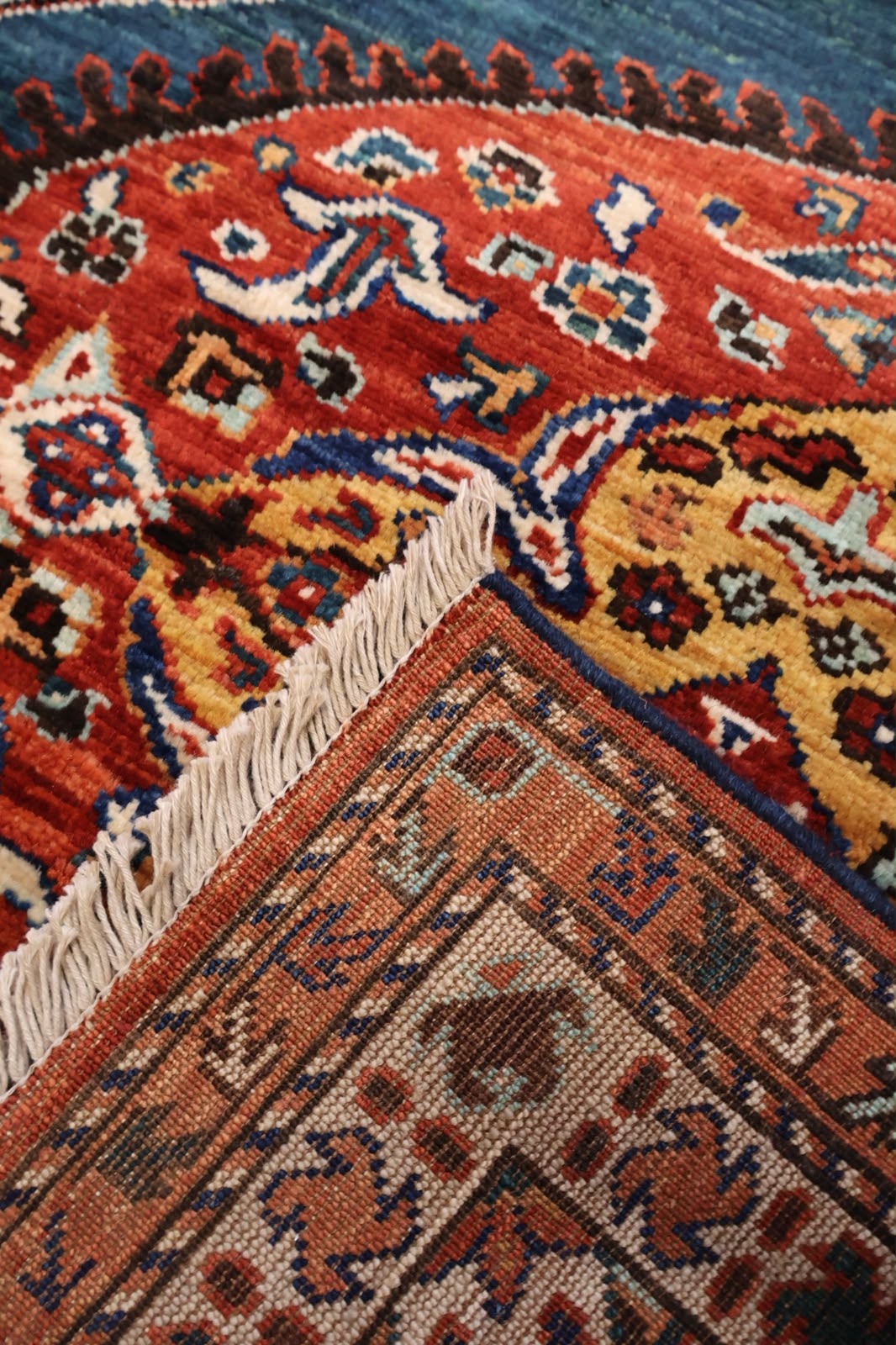 A close-up of a traditional patterned rug showing a corner with cream fringe, featuring red, orange, yellow, blue, and brown geometric and floral motifs.