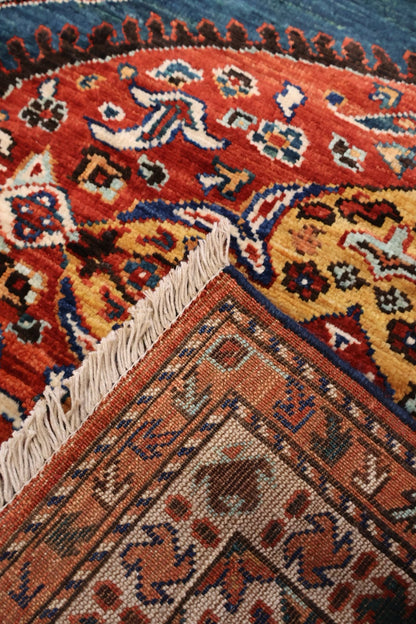 A close-up of a traditional patterned rug showing a corner with cream fringe, featuring red, orange, yellow, blue, and brown geometric and floral motifs.