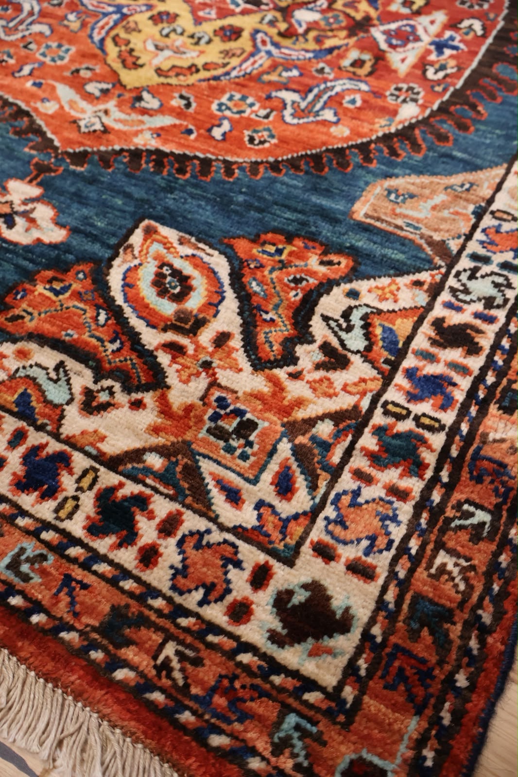 A close-up of a traditional patterned rug showing its detailed woven design, featuring a deep blue field, orange and red floral motifs, and cream borders with fringe.