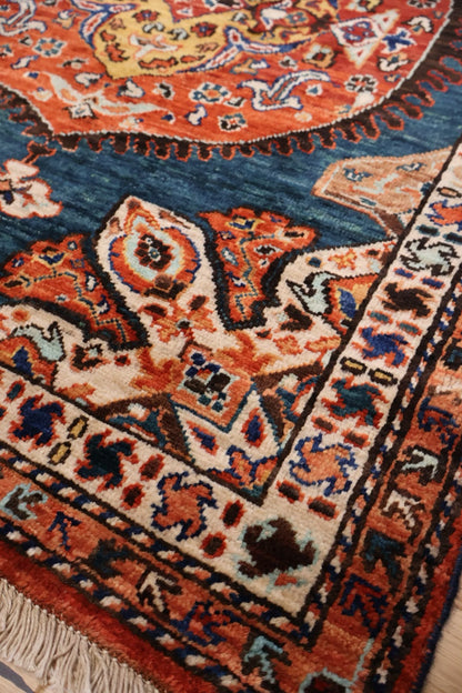 A close-up of a traditional patterned rug showing its detailed woven design, featuring a deep blue field, orange and red floral motifs, and cream borders with fringe.