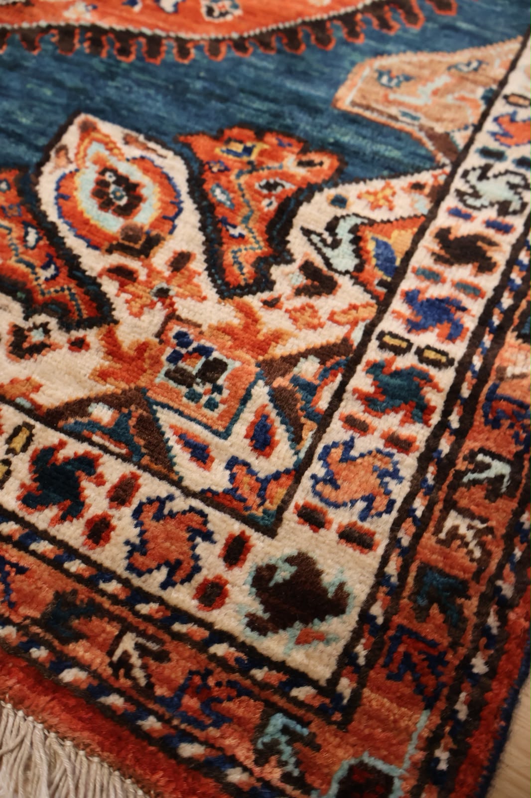Close-up of a traditional patterned rug corner showing cream, rust orange, blue, and black woven motifs with fringe visible along the edge.