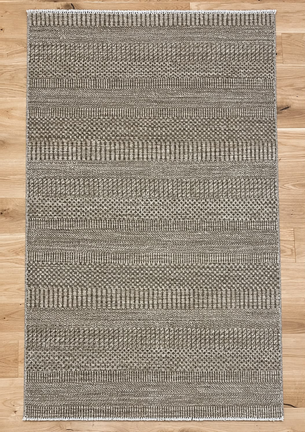 A rectangular woven area rug with a textured horizontal striped pattern in gray and beige tones, laid flat on a light wood floor.
