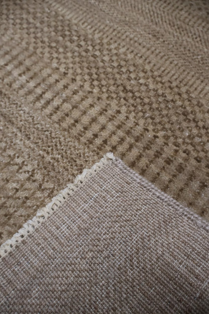 Close-up of a woven textile with a textured herringbone pattern in warm taupe and beige tones, showing the fabric weave and corner fold.