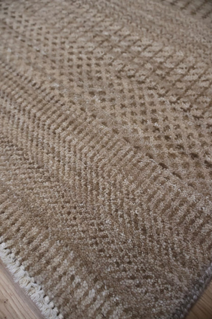 Close-up of a woven area rug with a textured basketweave pattern in beige and brown tones, showing the detailed pile and a small section of white fringe along the edge.