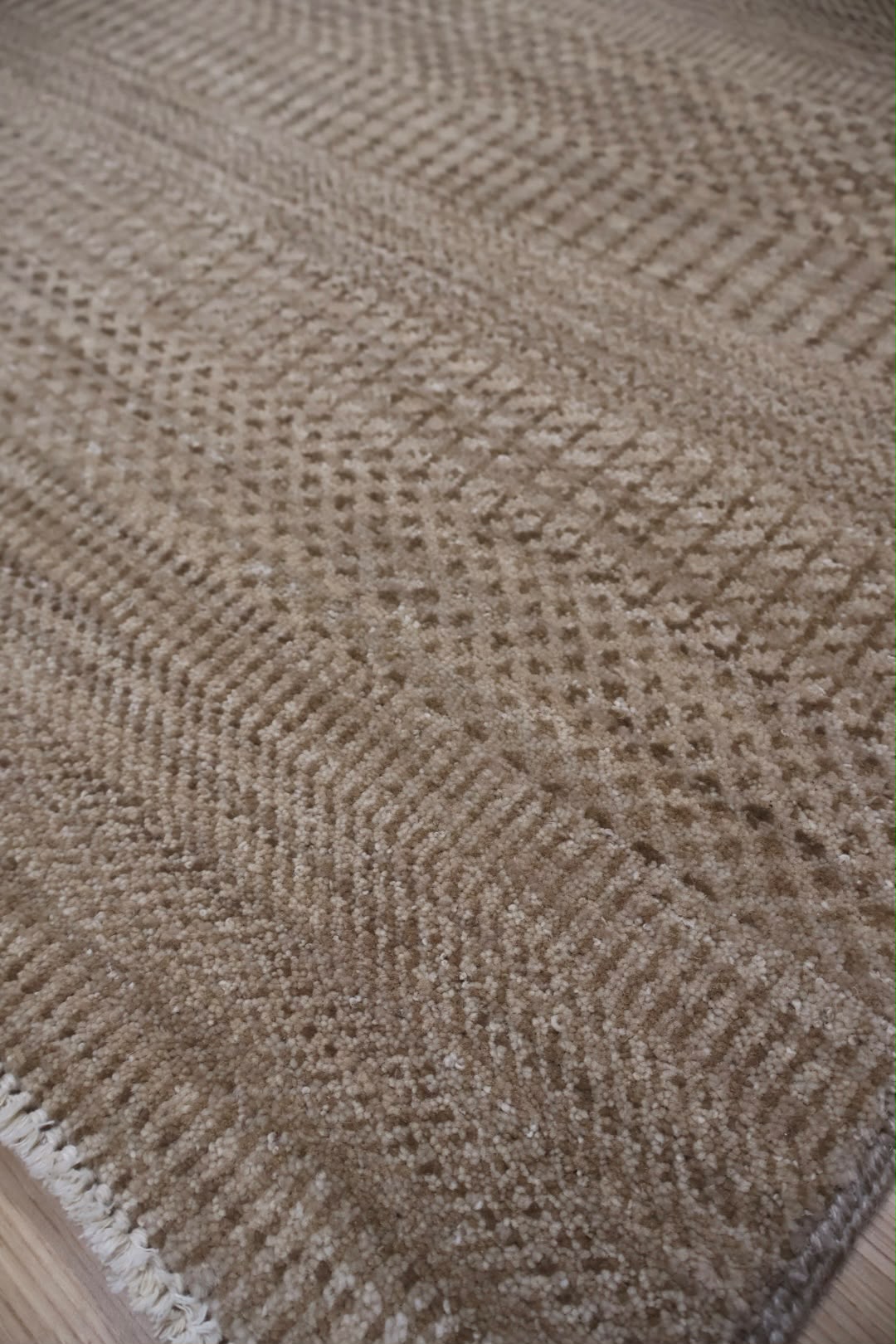 Close-up of a woven rug or textile with a textured herringbone pattern in warm beige and taupe tones, showing part of the edge and a hint of flooring underneath.