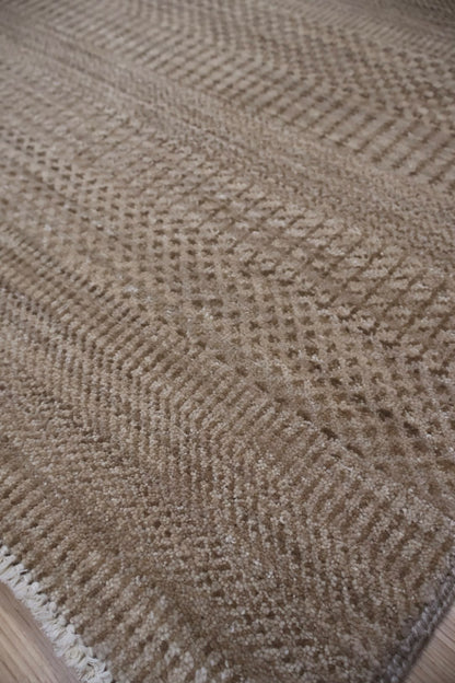 Close-up of a woven rug or textile with a textured herringbone pattern in warm beige and taupe tones, showing part of the edge and a hint of flooring underneath.