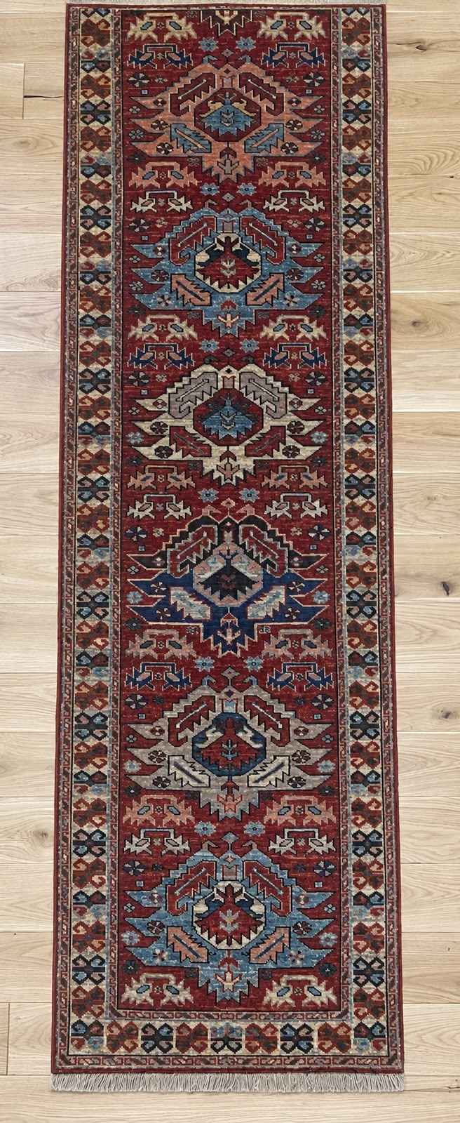A long rectangular area rug with an intricate traditional pattern in deep red, navy blue, beige, and brown, laid out on a light wood floor.
