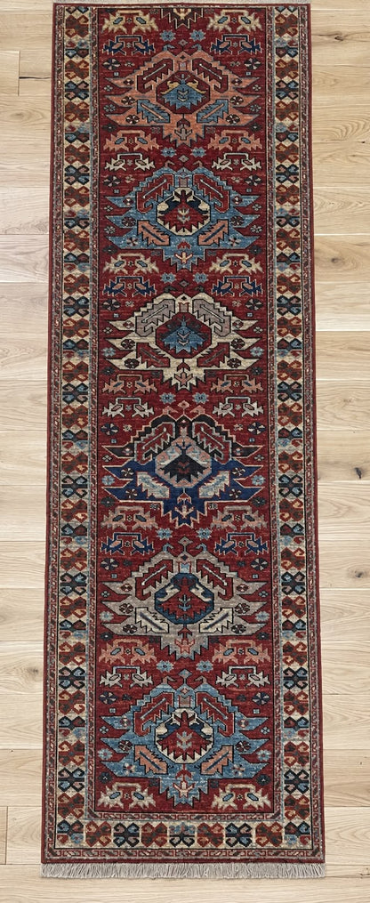 A long rectangular area rug with an intricate traditional pattern in deep red, navy blue, beige, and brown, laid out on a light wood floor.