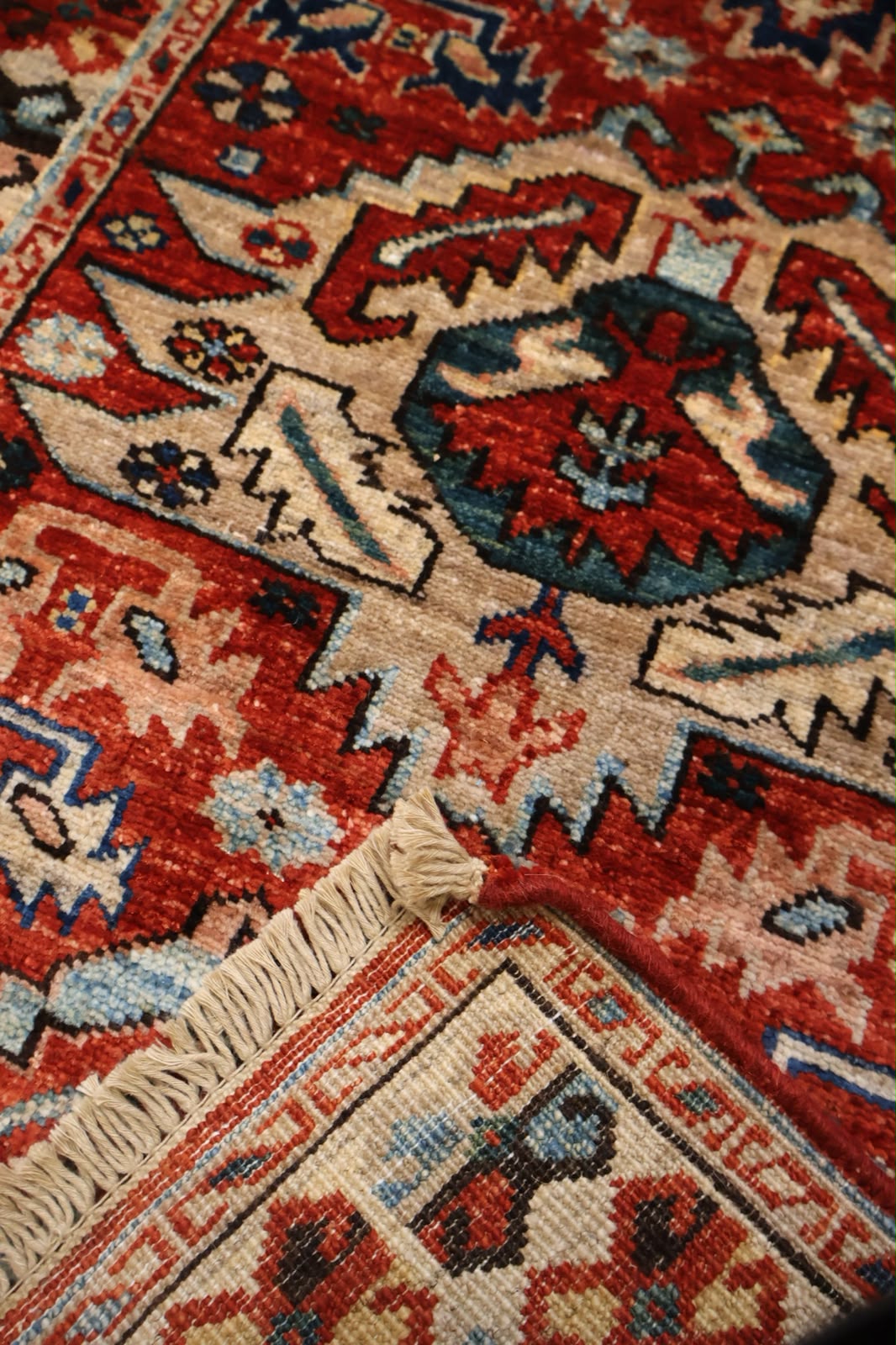 Close-up of a traditional patterned rug with red, beige, and blue geometric motifs and a cream fringe visible at the bottom left.