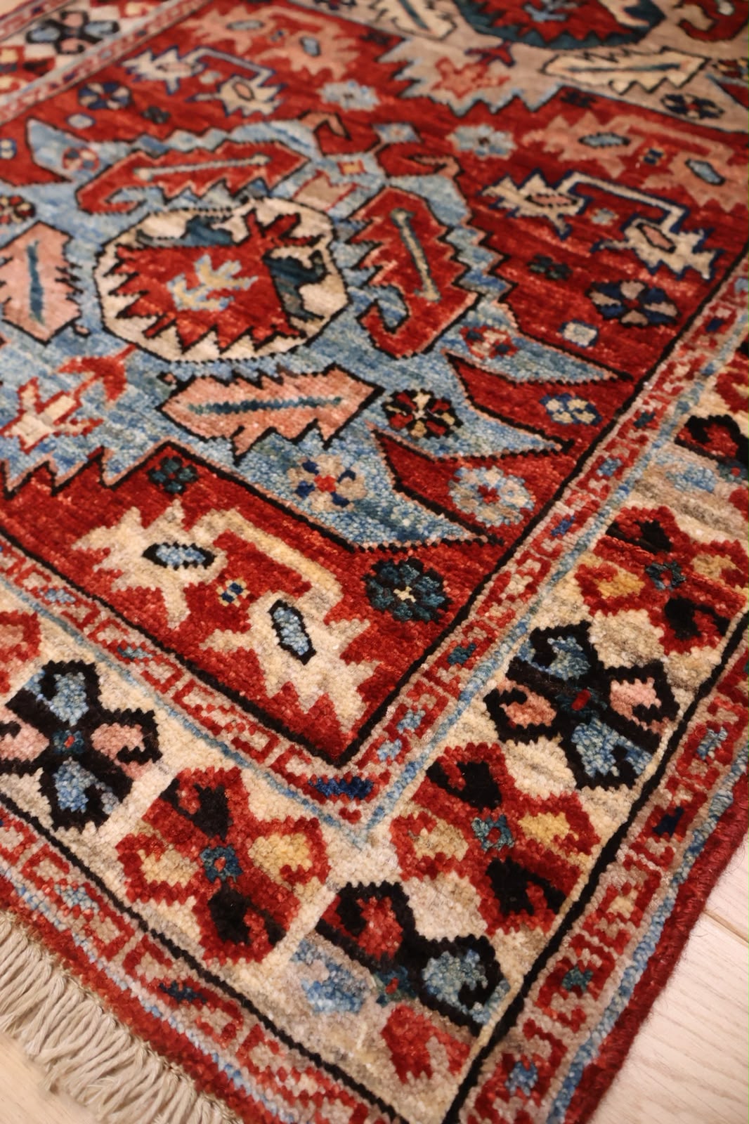 A close-up of a traditional handwoven area rug with a complex geometric pattern in red, beige, blue, and black, featuring a fringed edge on a light wood floor.